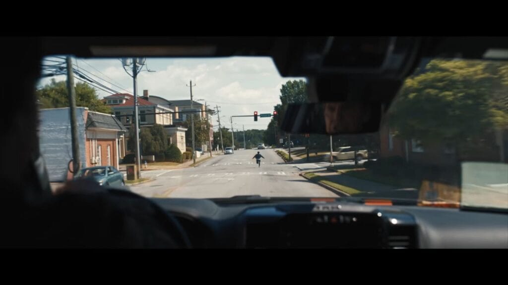 Point-of-view shot through a car windshield looking at a figure running directly toward the vehicle on a sunny road in Weapons.