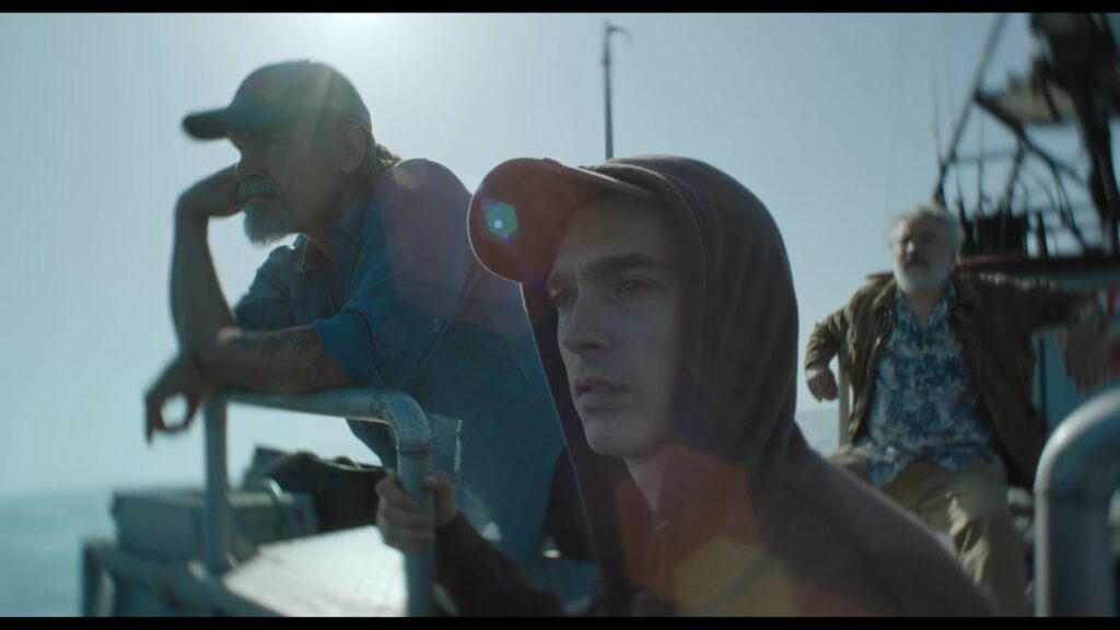 Austin Abrams wearing a hoodie leaning on a boat railing with older fishermen standing behind him under the sun in Whalefall.
