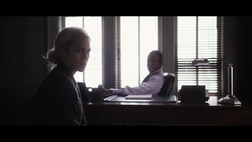 Brittany Snow sitting in an office looking off to the side with an interviewer in the background in Would You Rather.