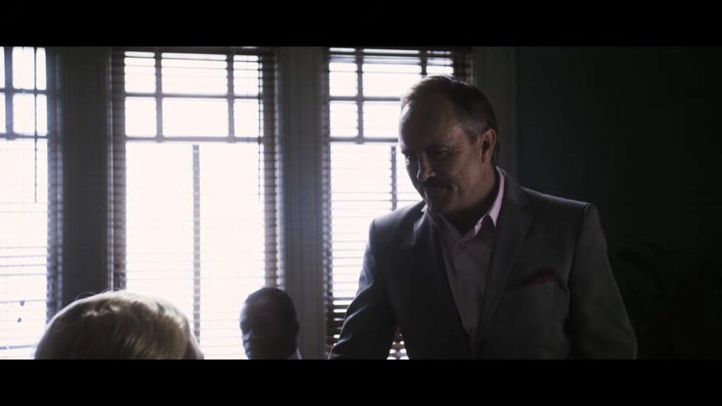 Jeffrey Combs standing in a suit and smiling menacingly in an office in Would You Rather.