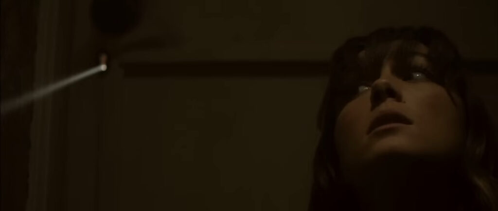 A low-angle close-up of Erin looking up nervously in the dark as a thin wire or string hangs nearby in You're Next.